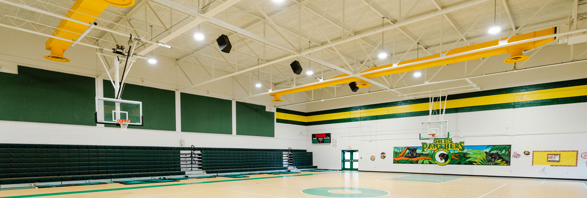 Gymnasium with basketball hoops, green bleachers and a green and yellow strip around the top of the wall at the Beaufort County Schools May River High School