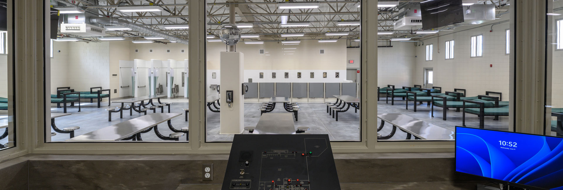 A jail dormitory with beds and metal tables and stools attached from viewpoint of the officer station at Volusia Jail built by Ajax Building Company.